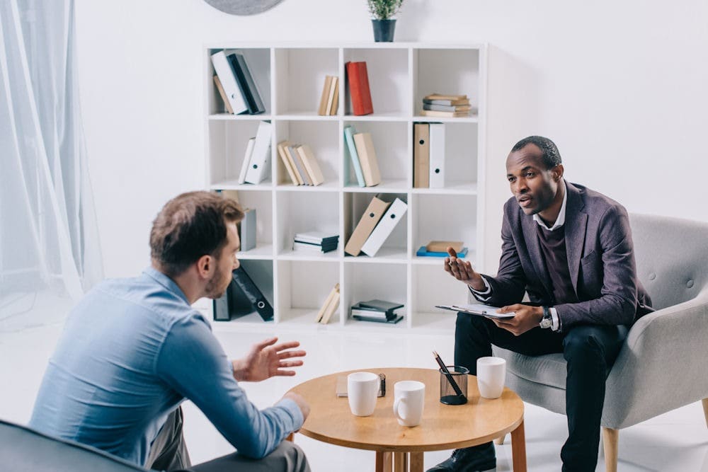 trigger-in-addiction Therapist in all-white office office explaining triggers in recovery to male patient asking questions