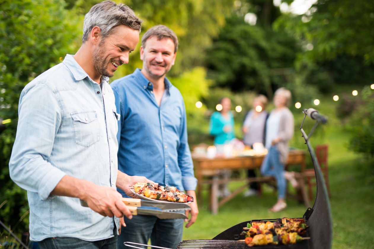 men prepares a barbecue for  friends gathered around a table in the garden